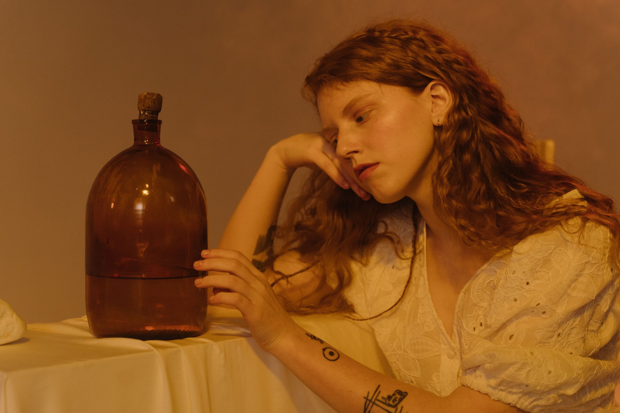 A woman resting her head on her hand beside a large amber jug on a white tablecloth